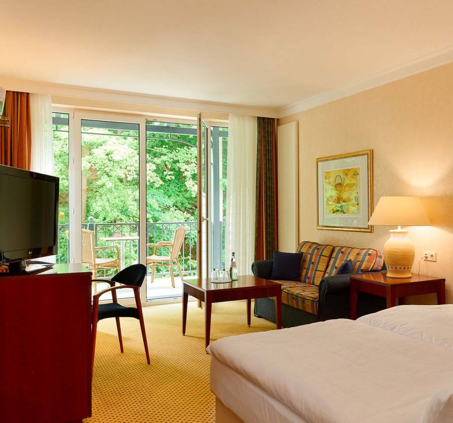 Granitz Deluxe Zimmer im Grand Hotel Binz mit großer Fensterfront, Sofa, Tisch und Balkon mit Blick ins Grüne.