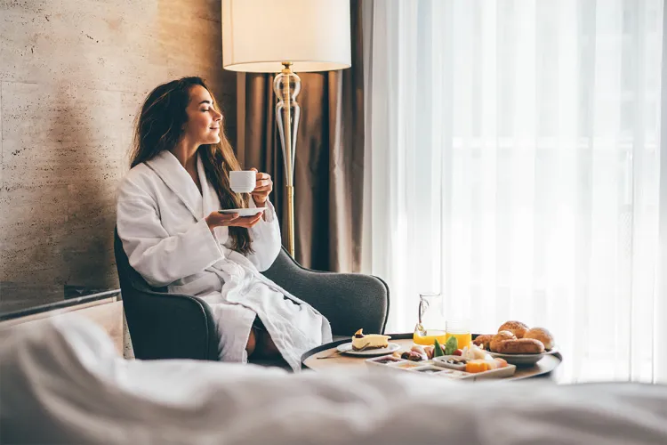 Frau im Bademantel genießt Kaffee beim Zimmerservice mit Frühstück im Hotelzimmer