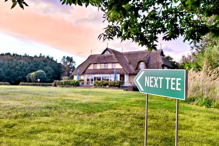 Außenansicht des Golfcentrums bei Sonnenuntergang. Im Vordergrund verweist ein Schild auf den nächsten Abschlag.