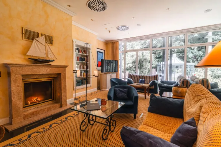 Das gemütliche Kaminzimmer des Grand Hotel Binz mit bequemen Sesseln, Kamin und Blick ins Grüne.