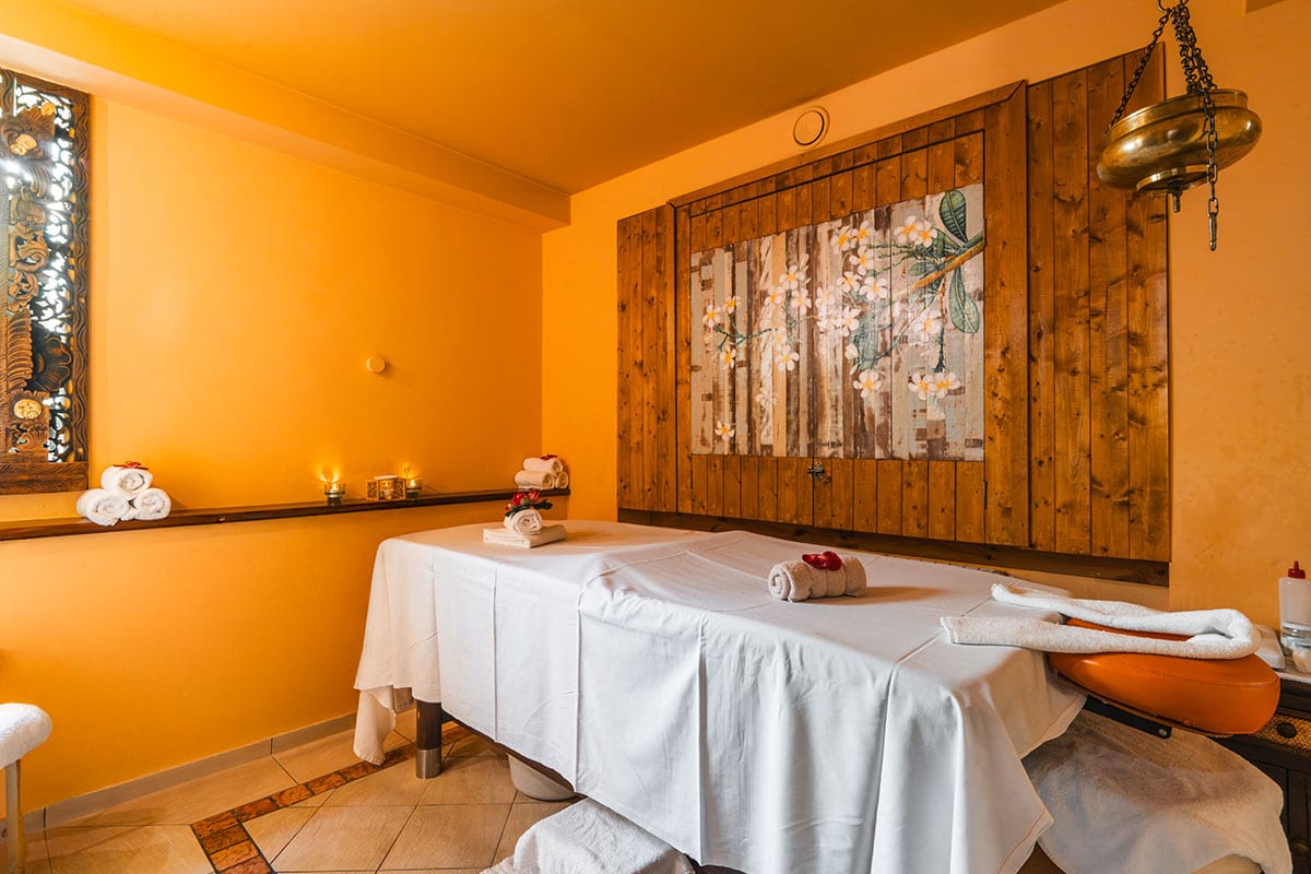 Massageraum mit weiß bezogener Liege, Kerzen, Holzdekor und Blüten im Spa